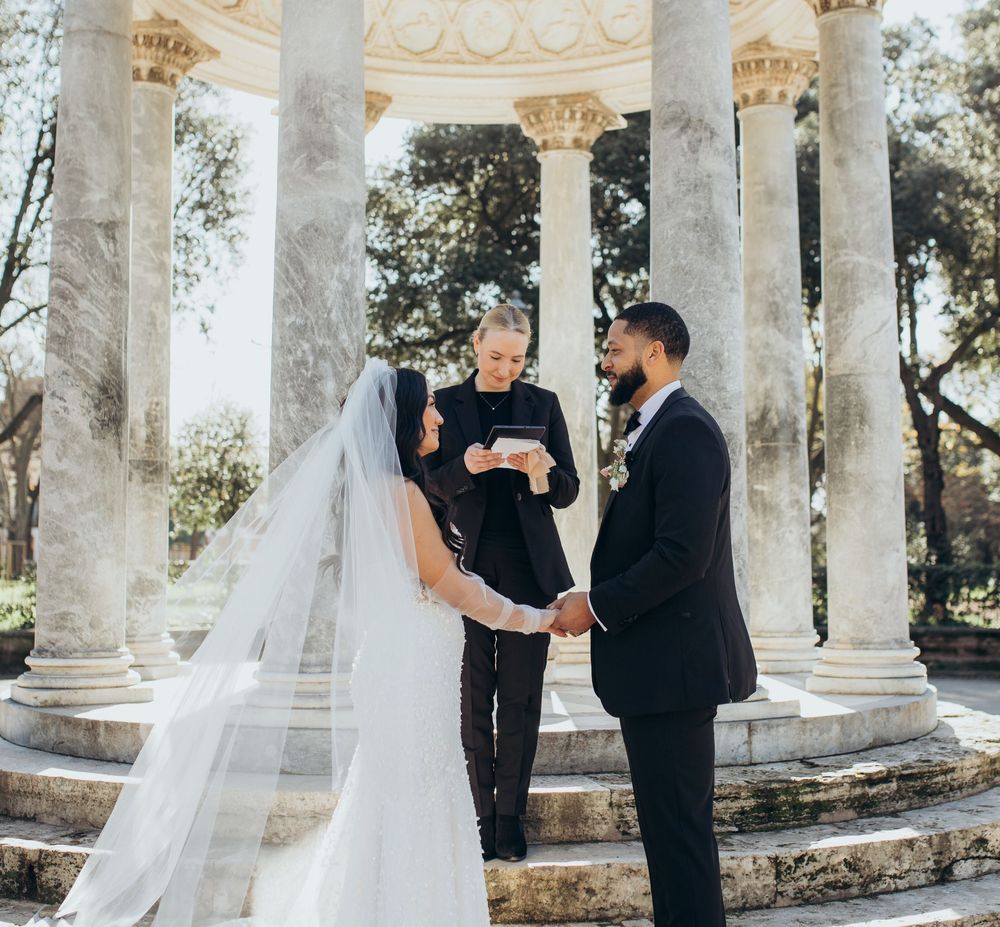 A wedding ceremony conducted under an ornate white marble pavilion with Corinthian columns in a garden setting.