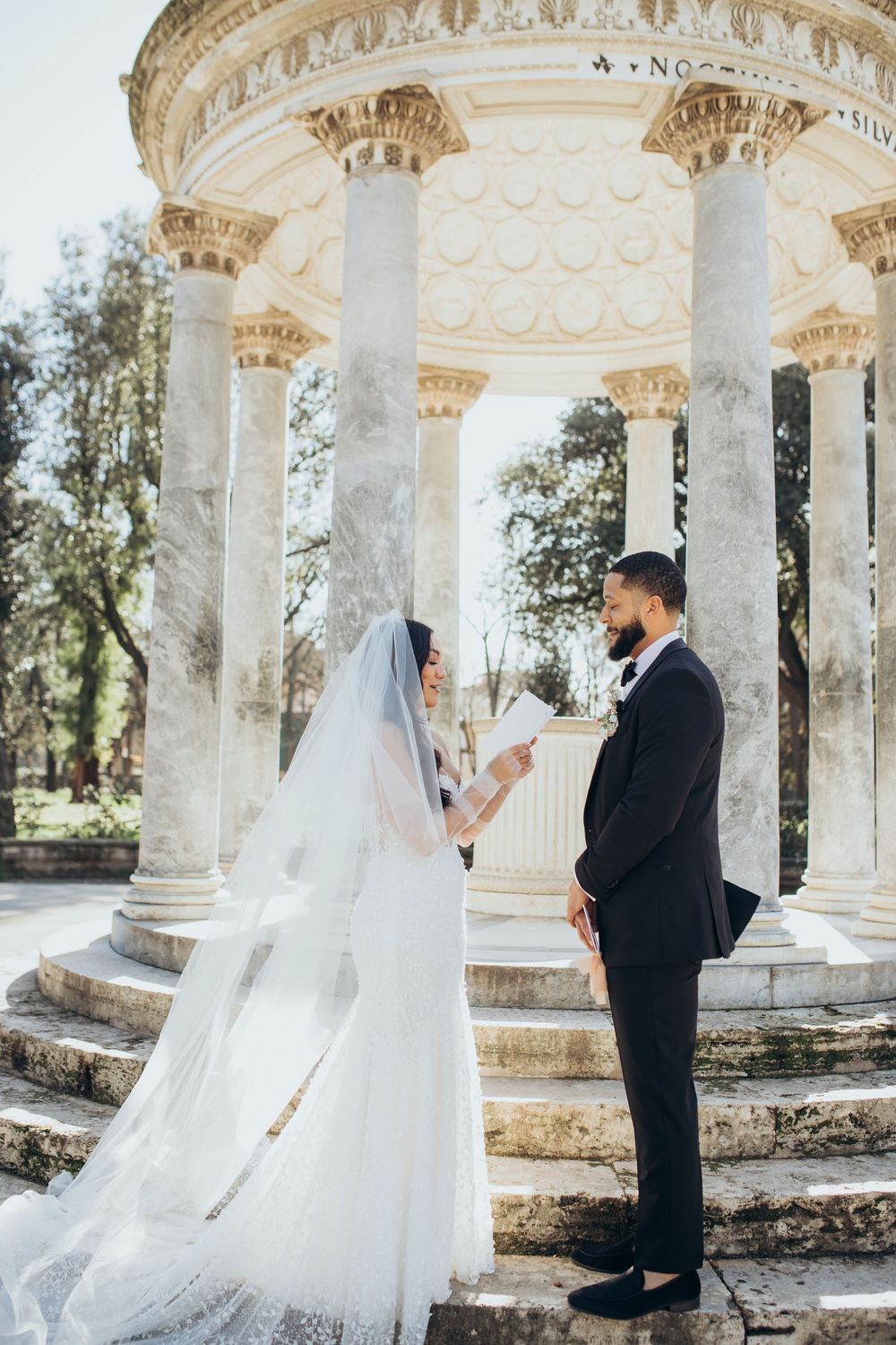 A couple exchanges vows beneath an ornate white marble gazebo surrounded by trees during their wedding ceremony.