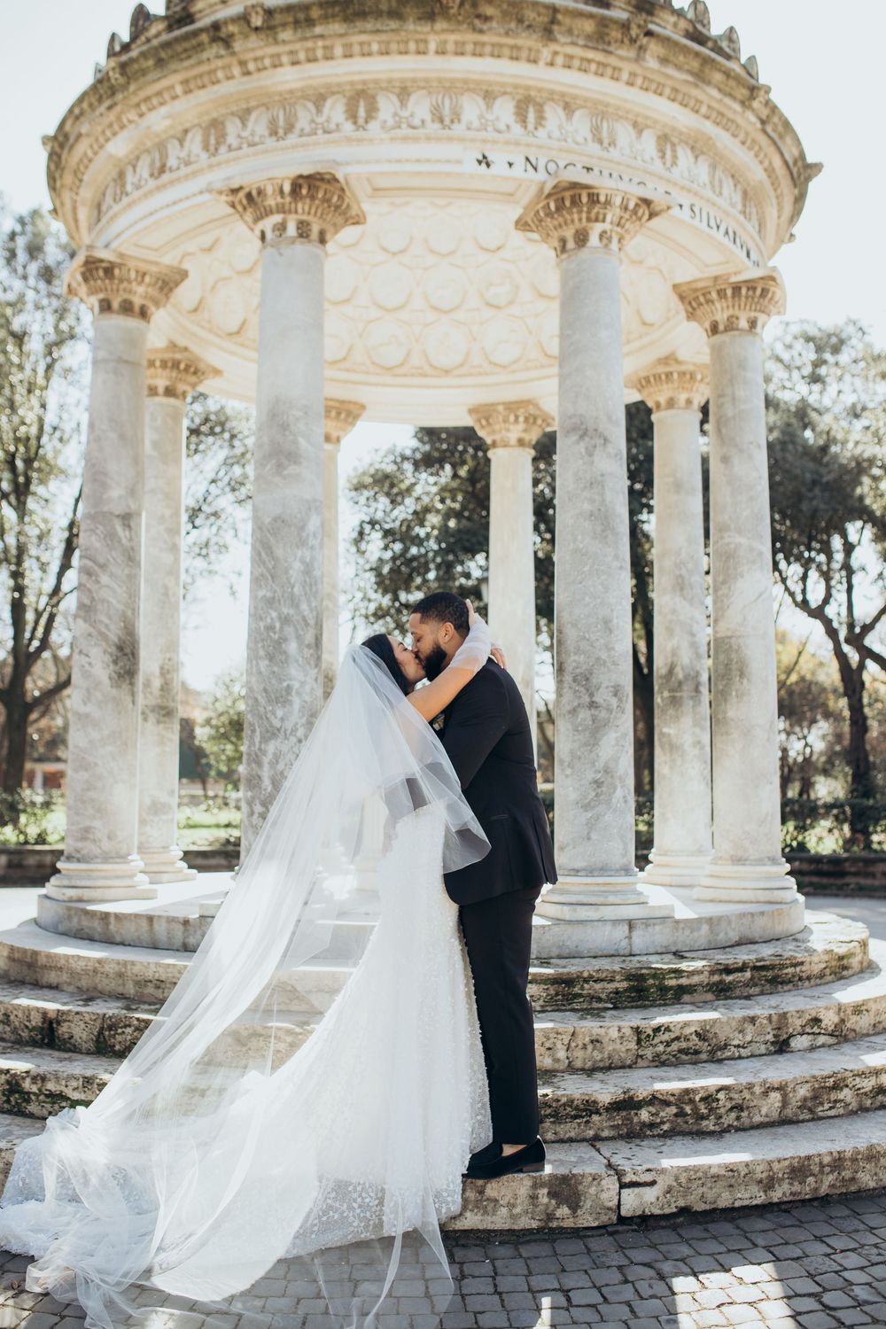 A couple embraces under an ornate white marble gazebo in a garden setting.