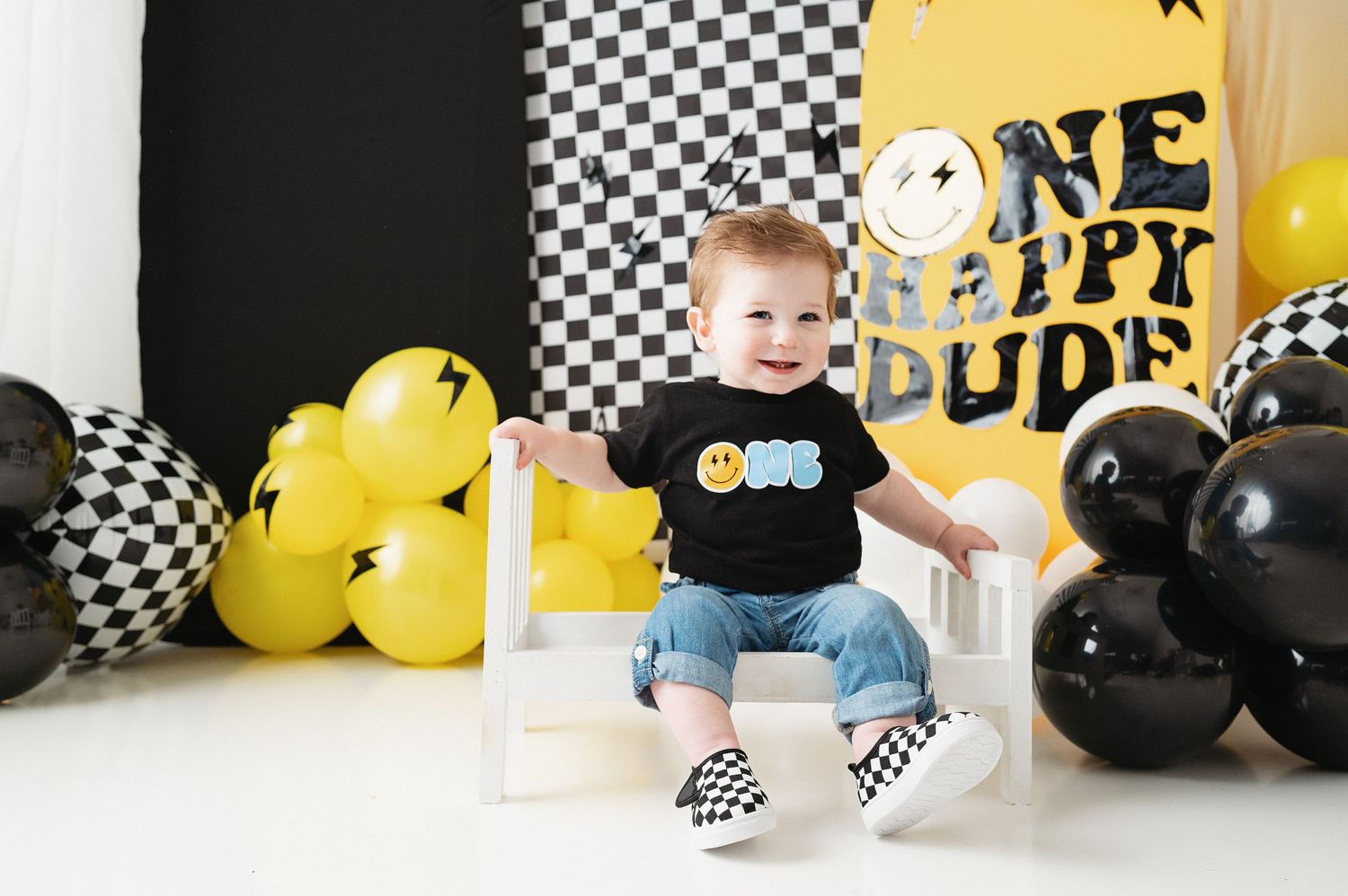 Child in black ONE shirt sitting on white chair with checkered shoes and balloon decorations.