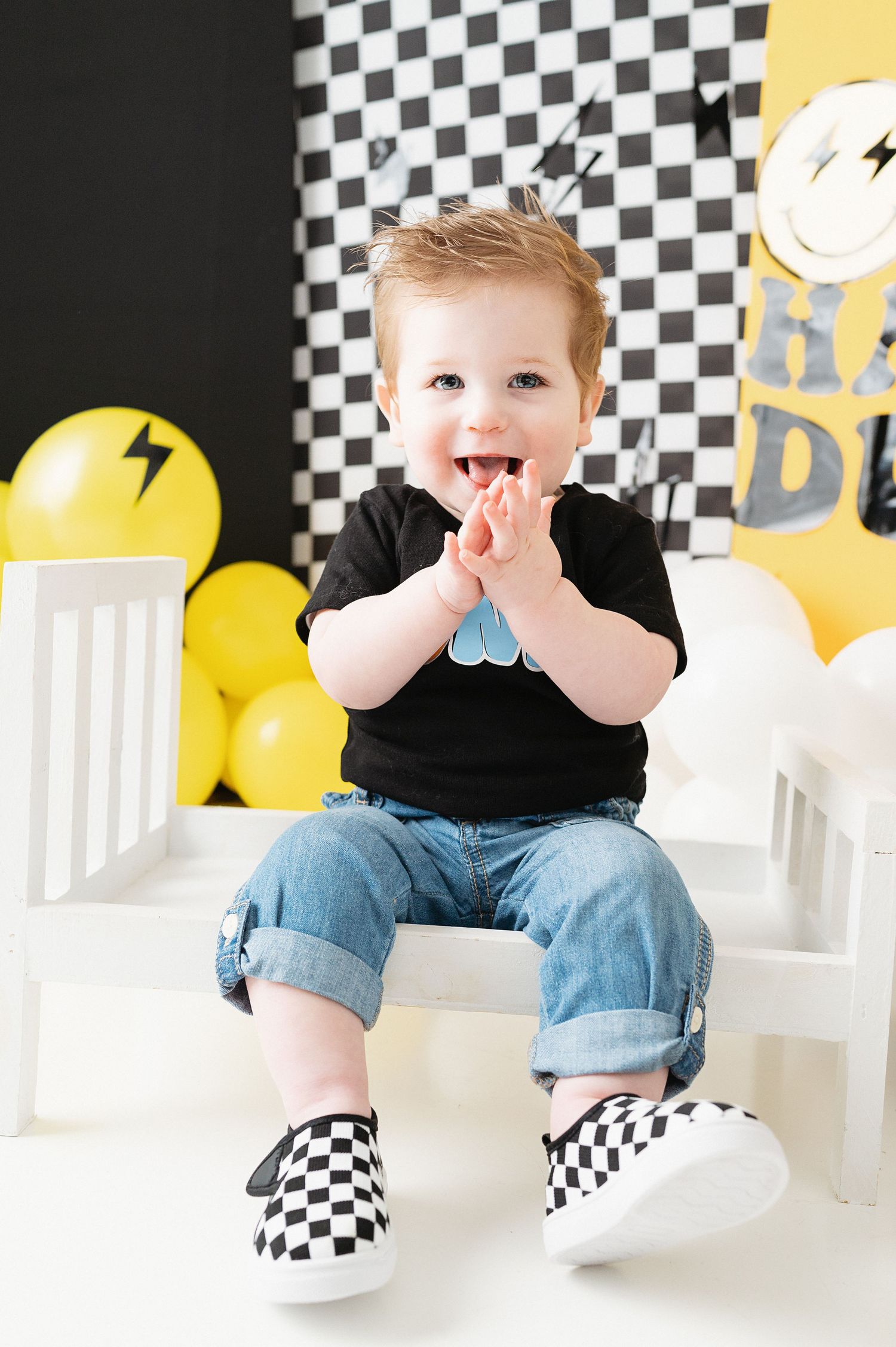 Toddler in checkered shoes sitting on white chair clapping against party backdrop.