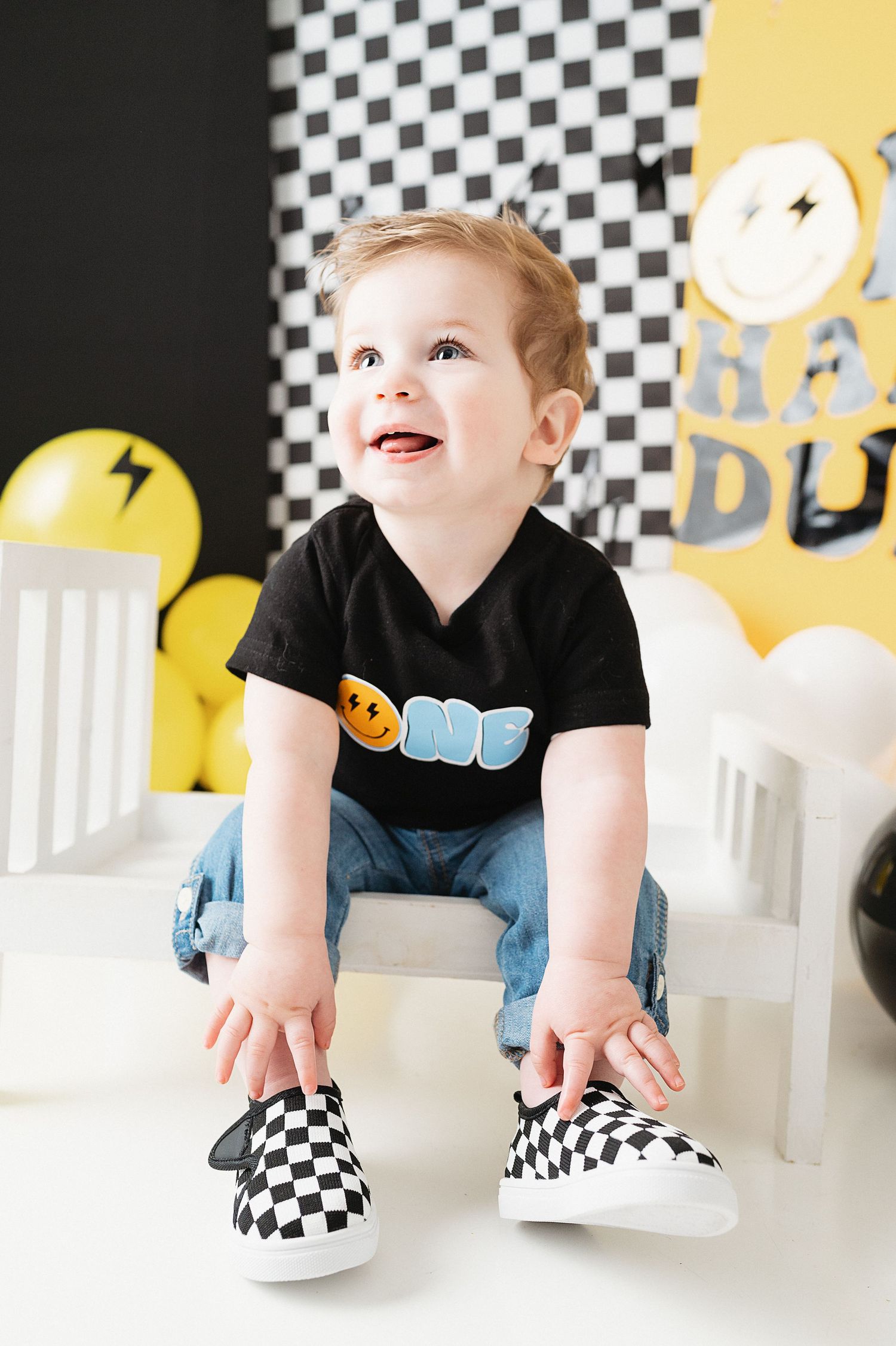 Child in ONE shirt sitting on white chair with checkered slip-on shoes.