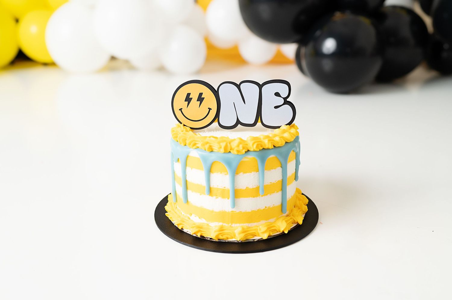 Yellow and blue drip cake decorated with smiley face ONE topper and black base.