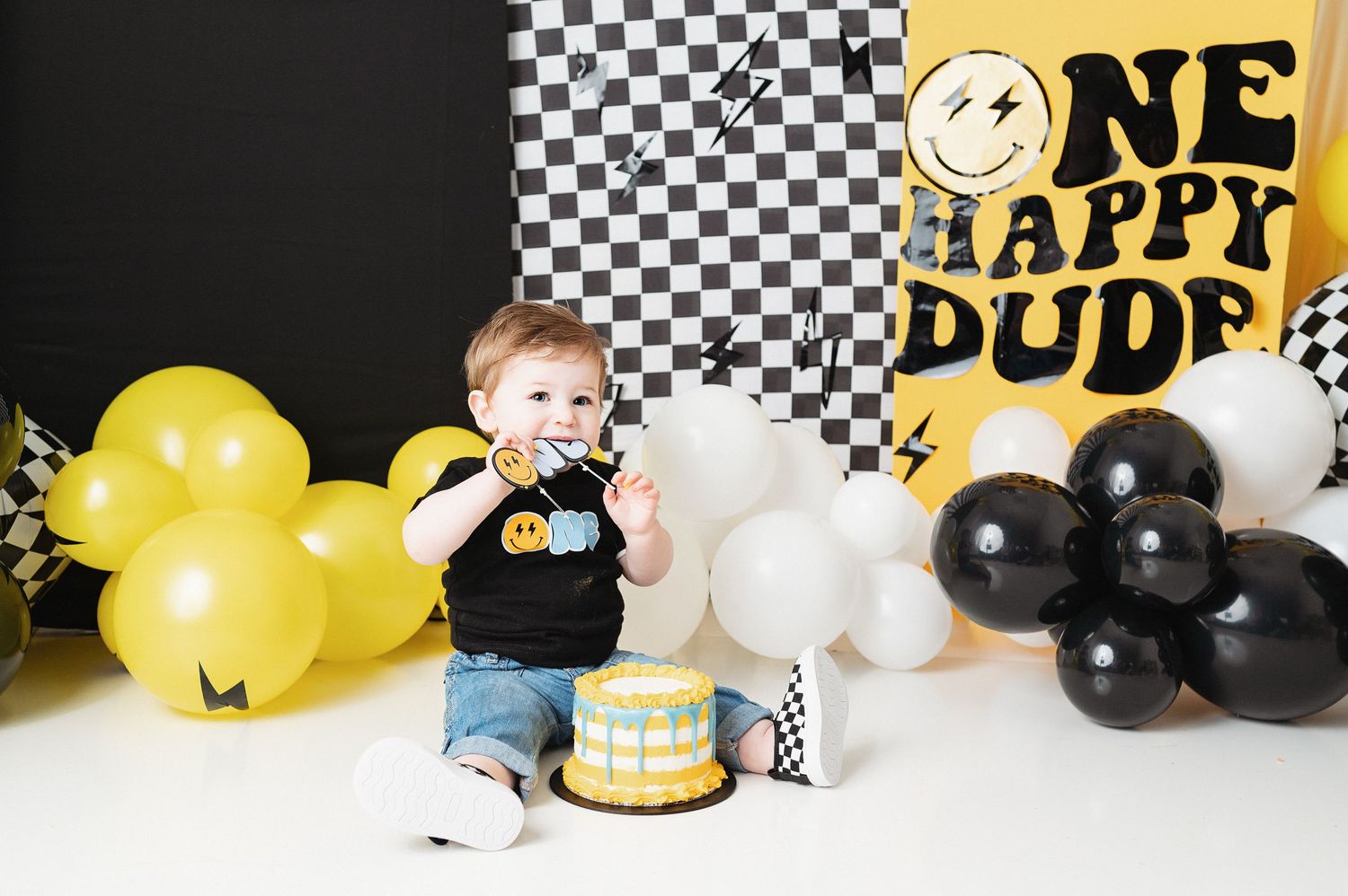 Smash cake photo setup with checkered backdrop, yellow and black balloons, and small cake.