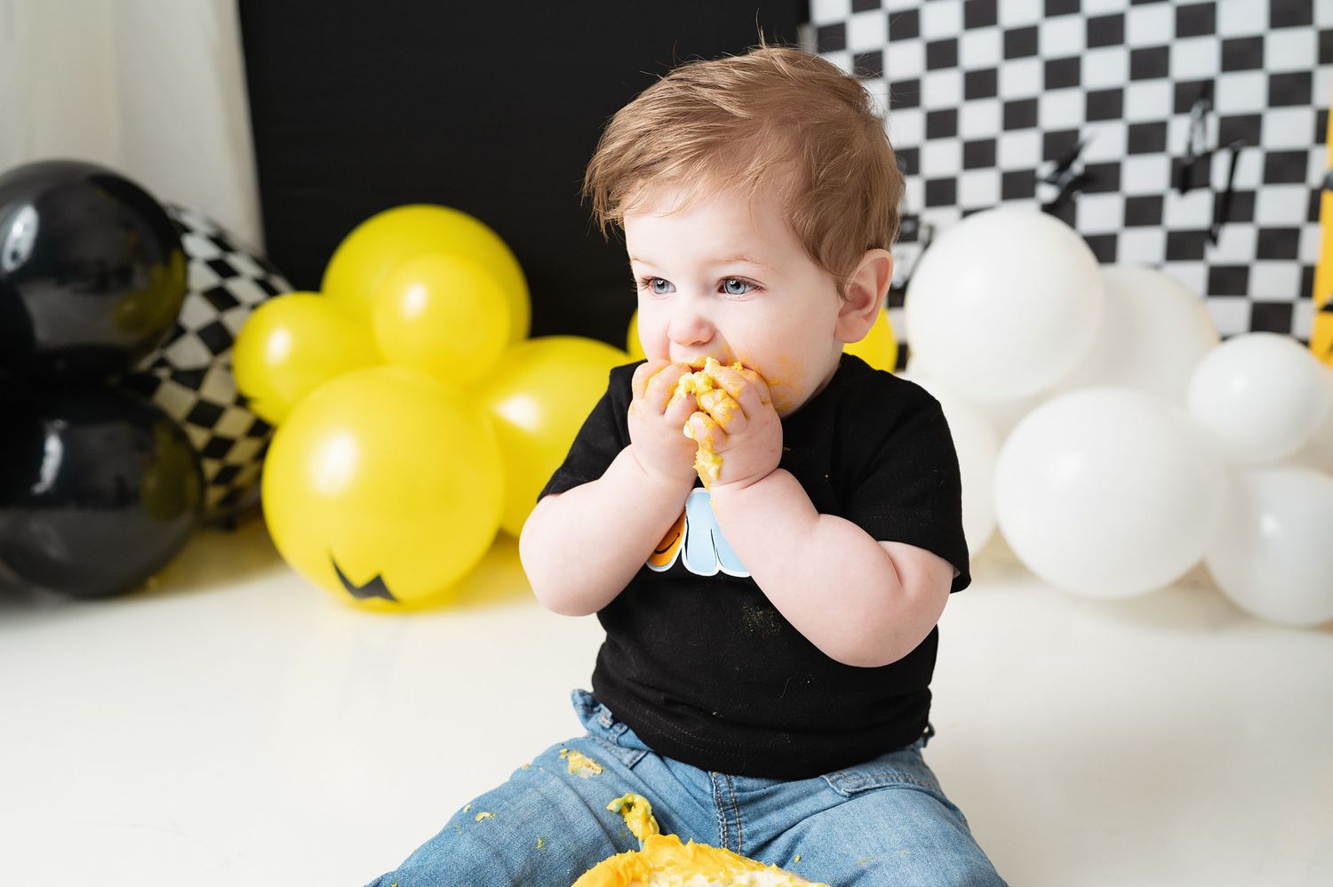 Child in black shirt enjoying cake against backdrop with yellow and black balloons.