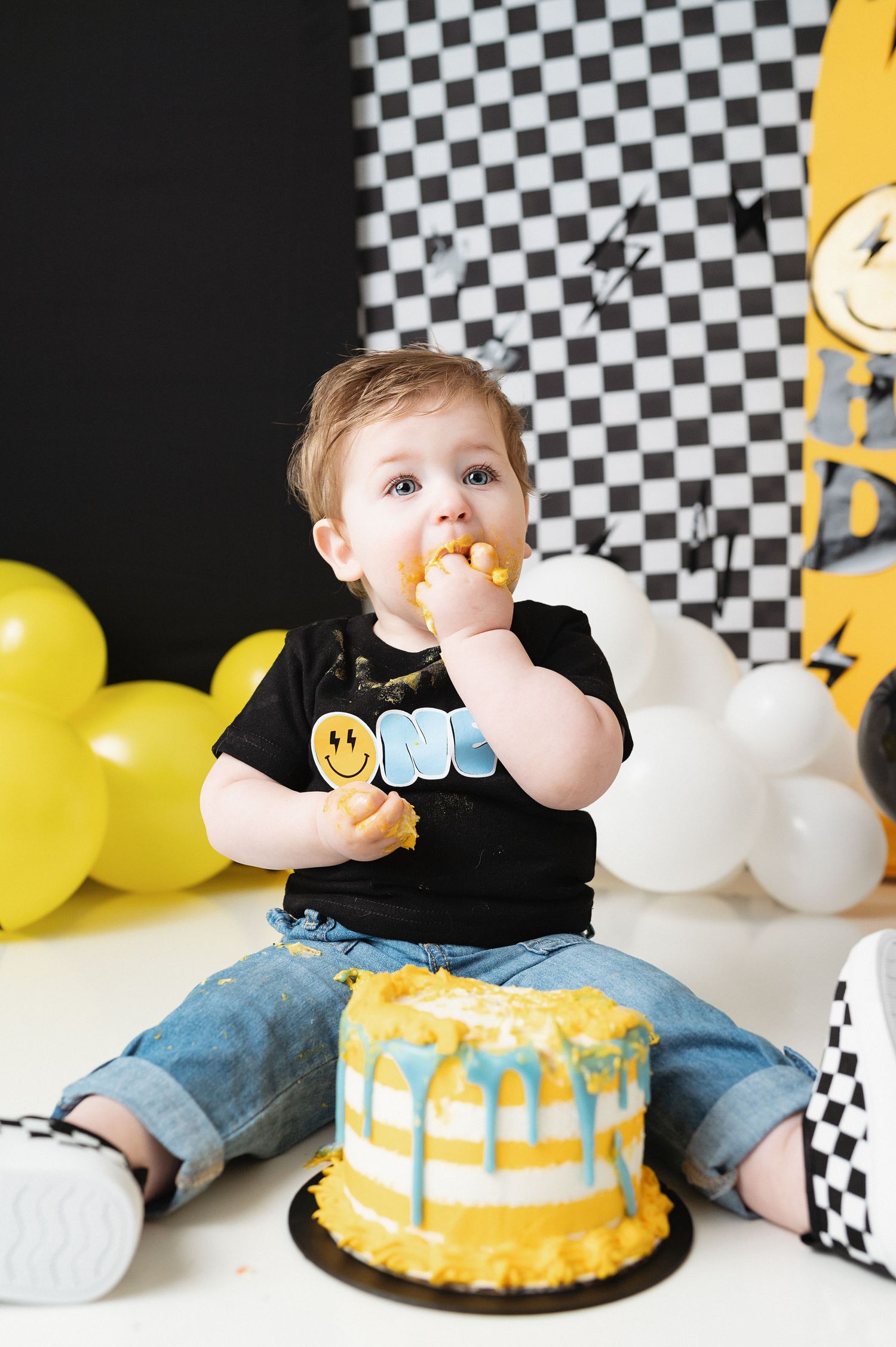 Baby in ONE shirt tasting yellow birthday cake with checkered and balloon decorations.