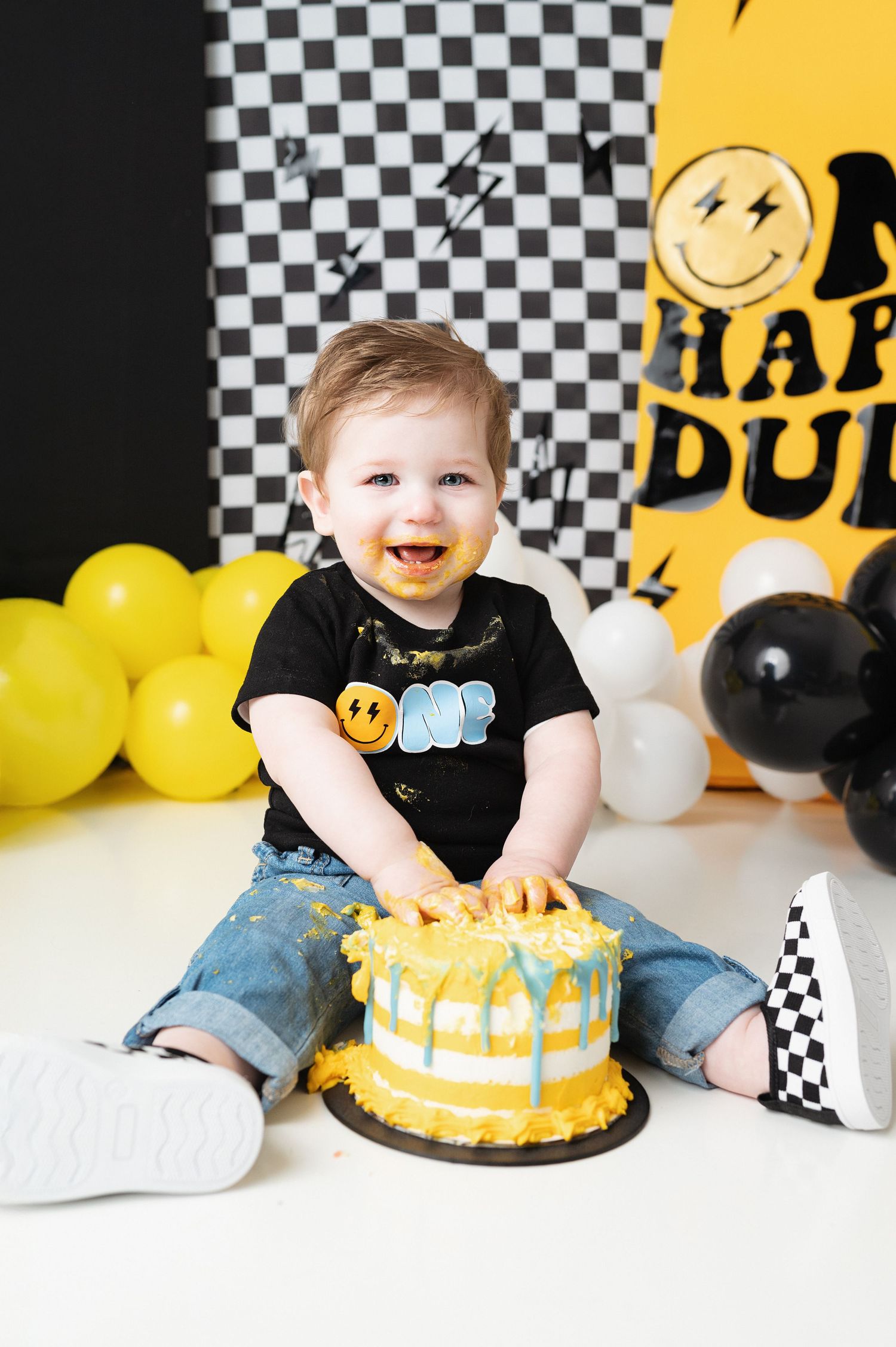 First birthday celebration with smiling baby and yellow drip cake against checkered backdrop.