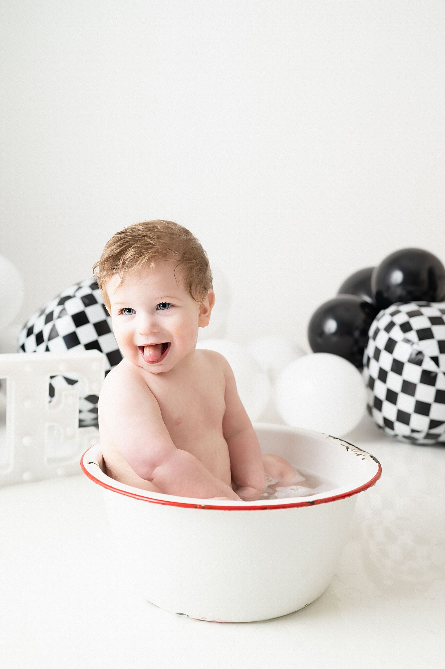 Baby laughing while sitting in white enamel basin with checkerboard balloons.