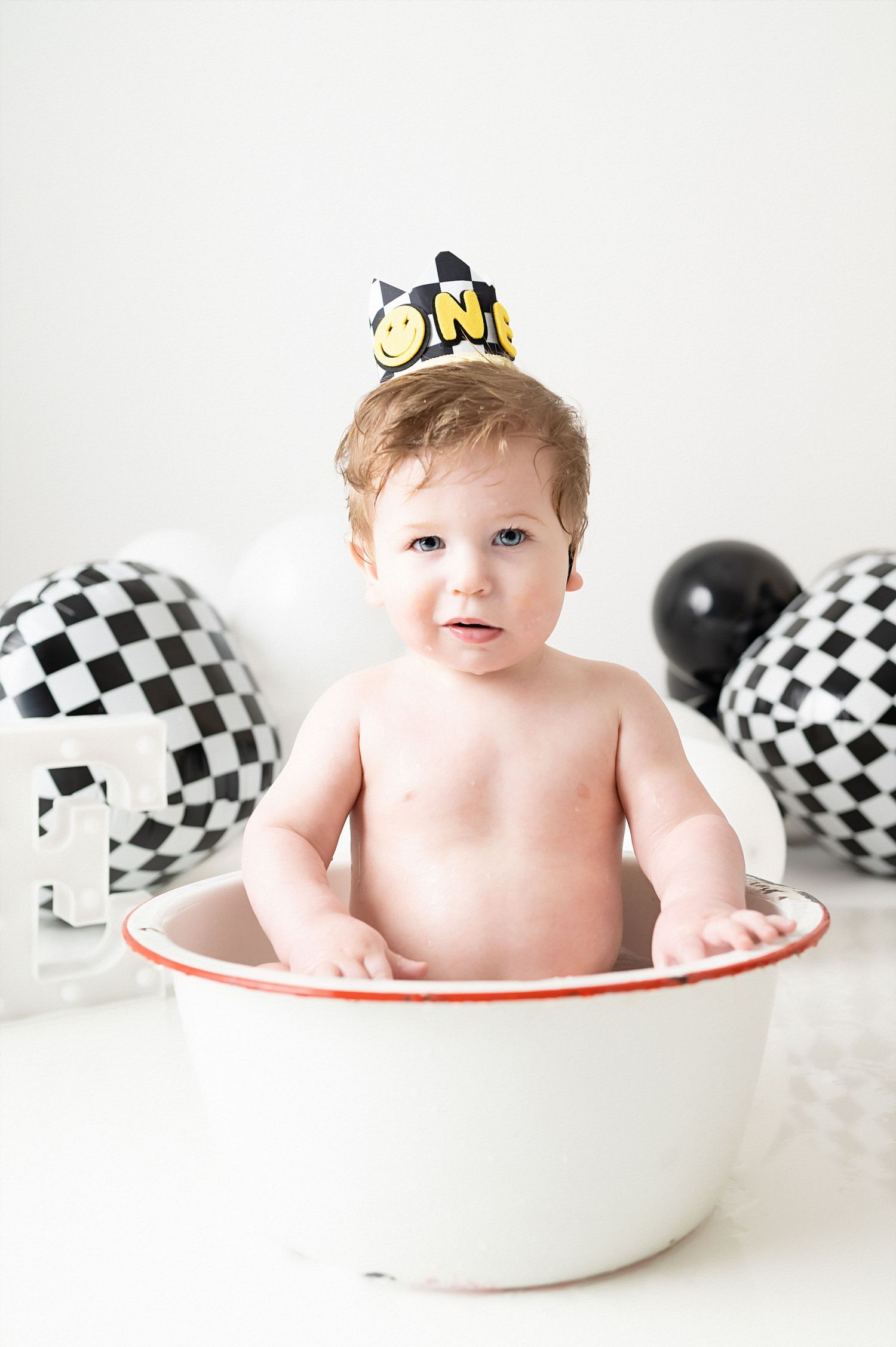 Baby sitting in white enamel bowl with checkered balloons and party crown for first birthday photo shoot.