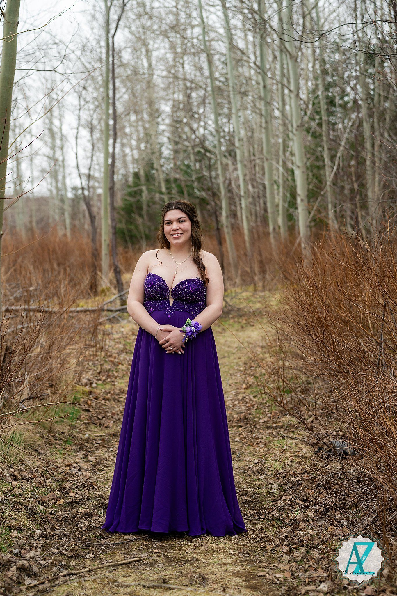 Person wearing a purple formal evening gown standing in a wooded area with bare trees and brown grass in winter.