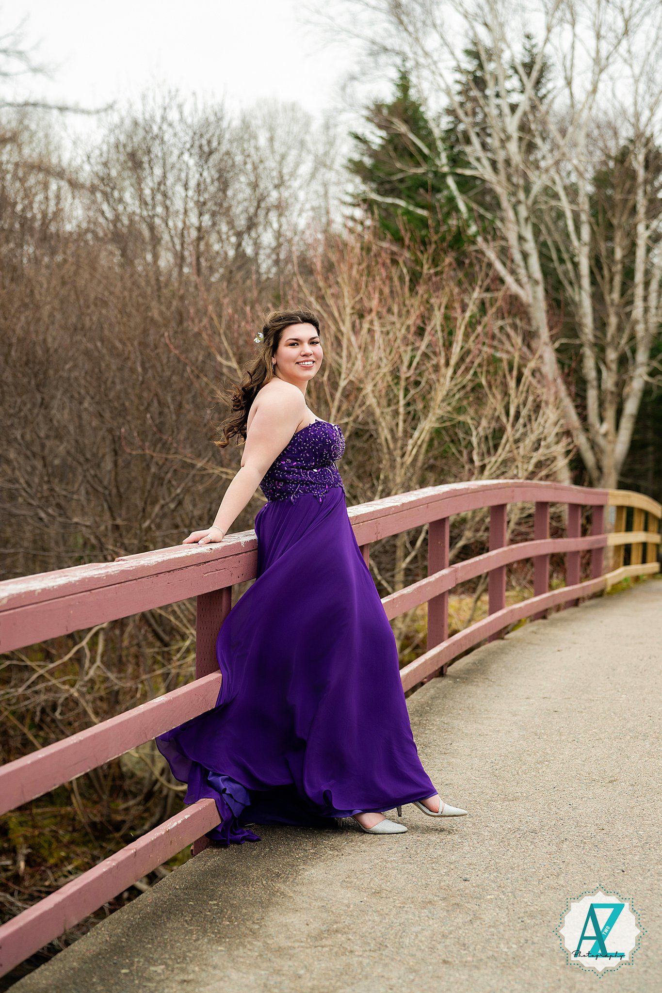A flowing purple evening gown sweeps elegantly across a wooden bridge during a spring photoshoot.