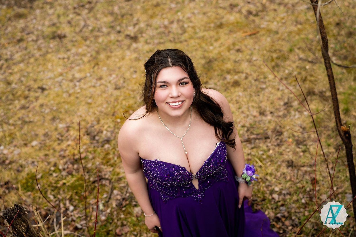 A young woman in a sparkly purple formal dress poses outdoors against a natural autumn background.