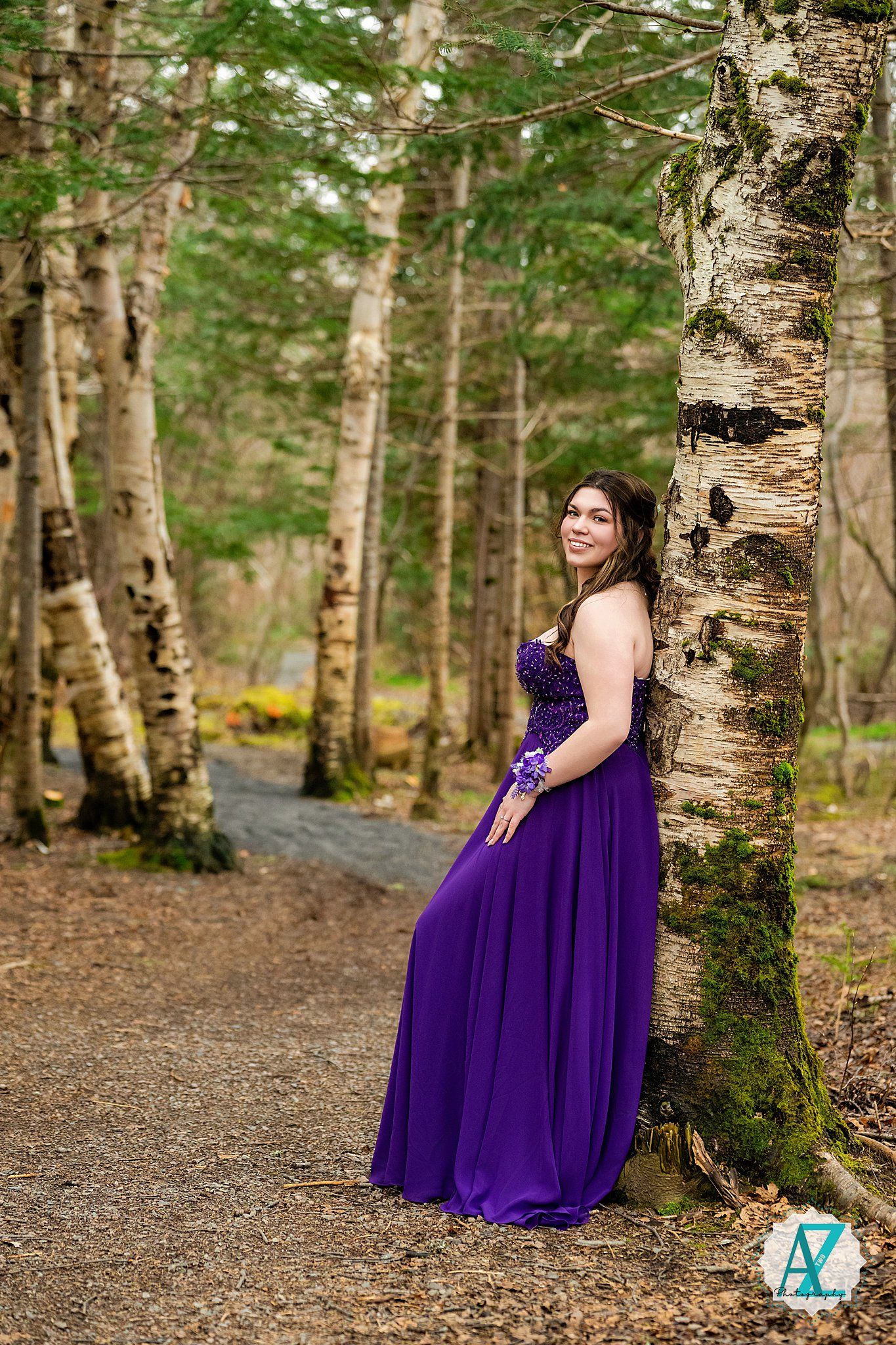 A woman in a flowing purple formal gown poses along a tree-lined forest path on a spring day.
