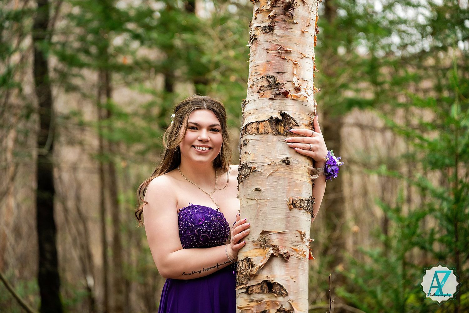 Someone in a purple formal dress poses next to a white birch tree trunk in a forest setting.