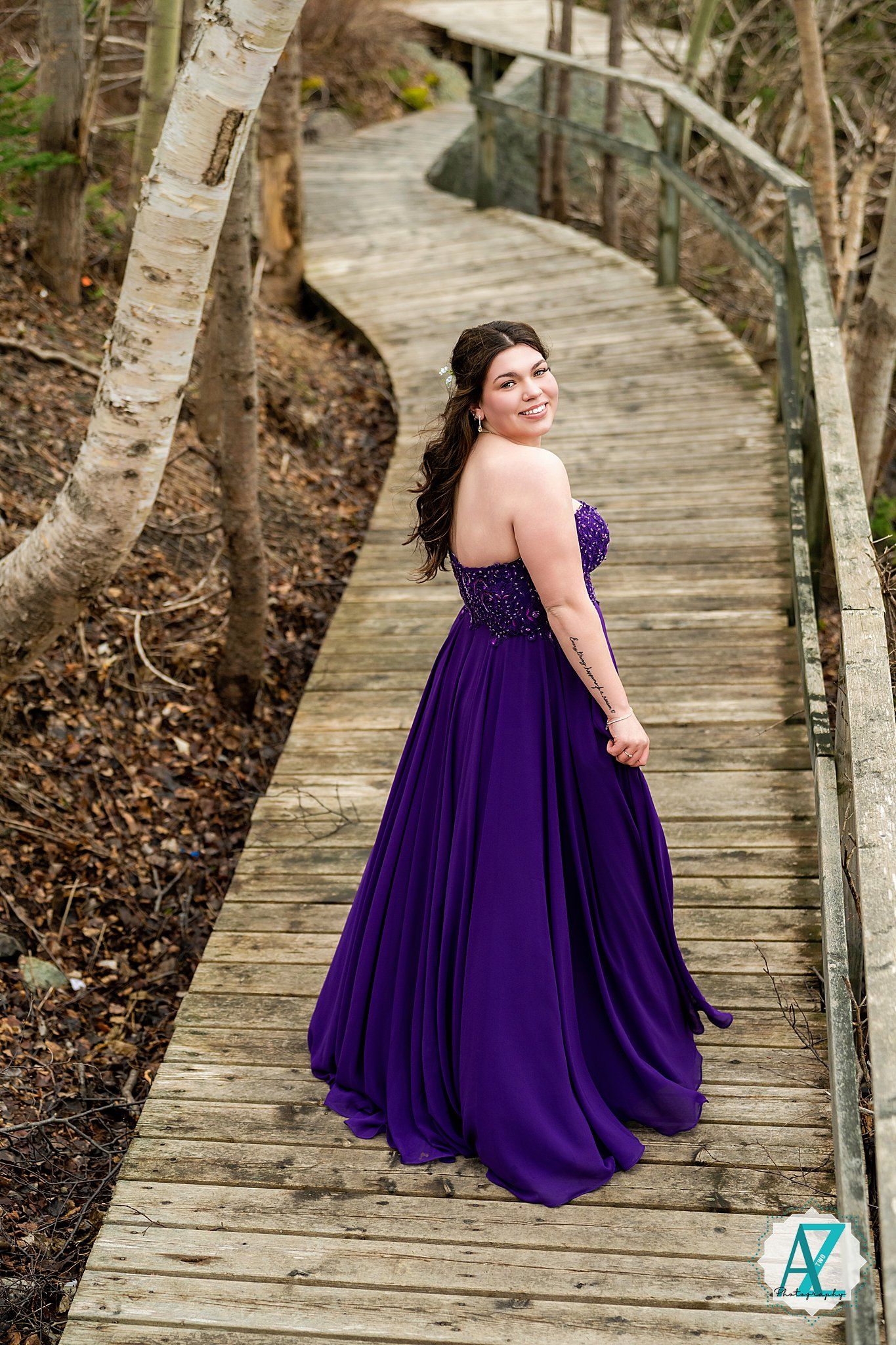A strapless purple formal gown glimmers in the light as the wearer stands on a winding wooden boardwalk path through trees.