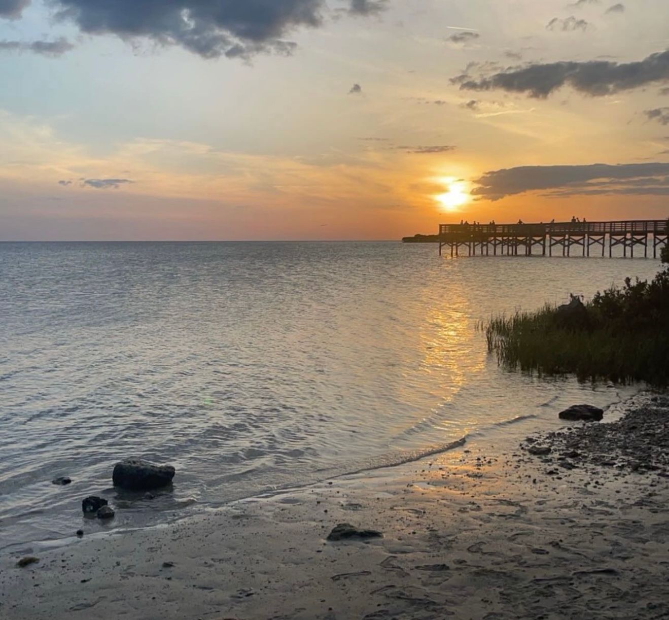 Panoramic view of a coastal sunset over calm waters with silhouetted pier and marsh grasses along the shoreline.