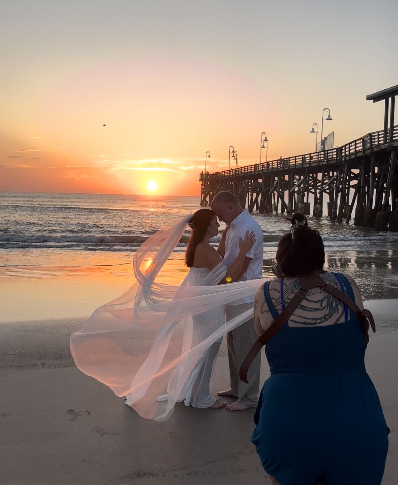 A photographer captures a couple standing near the ocean waves during a romantic beach sunrise photoshoot.