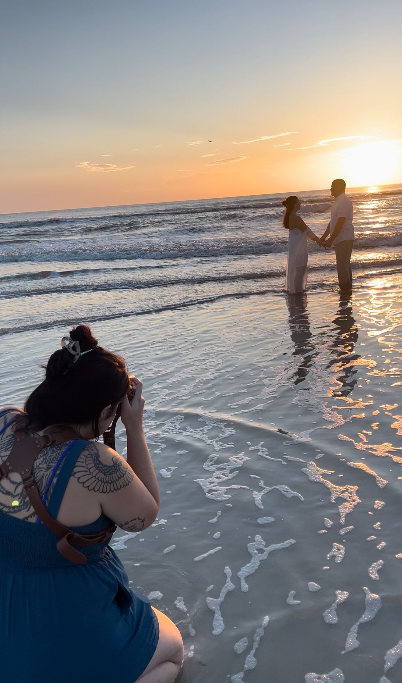 A photographer captures a couple standing in the ocean waves during a romantic beach sunrise photoshoot.