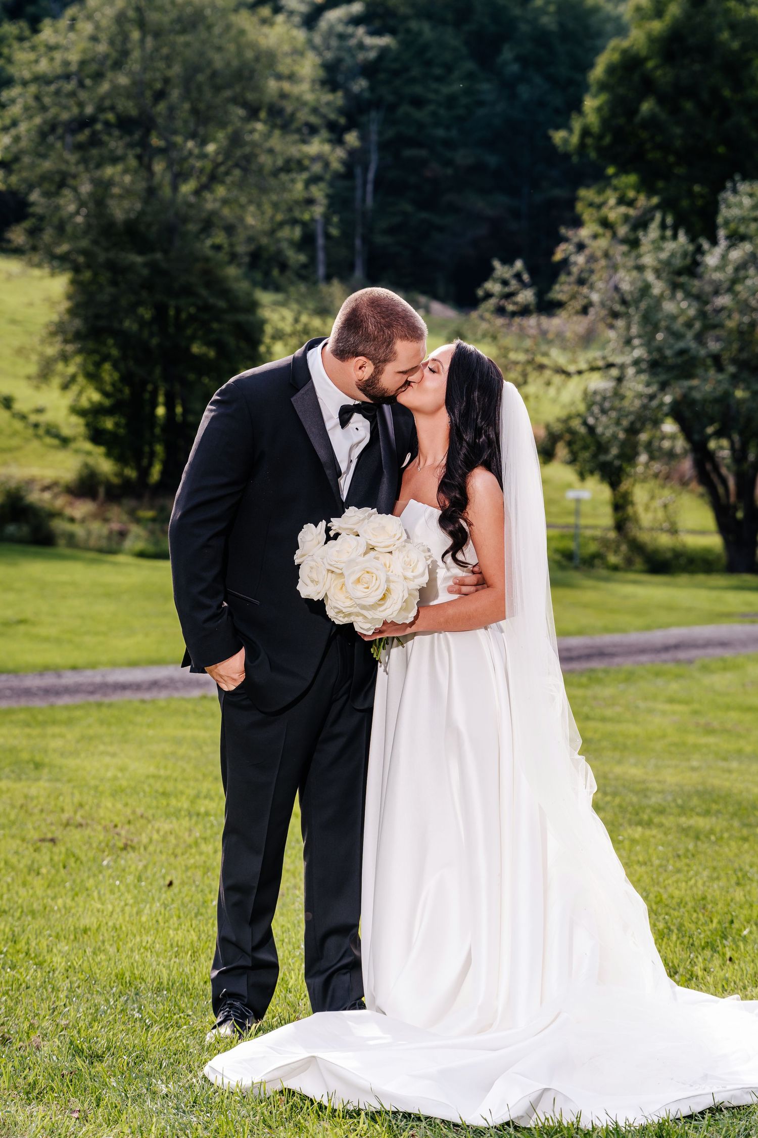 Couple in wedding attire share intimate moment outdoors on green lawn with white bouquet and flowing bridal veil.