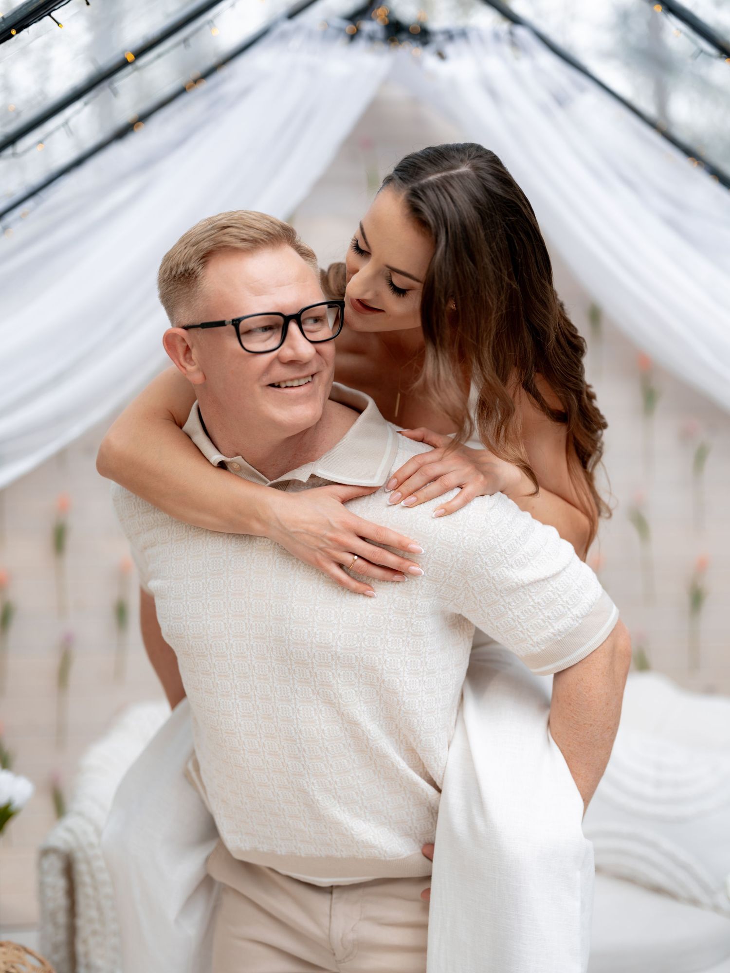 A romantic couple embraces in a dreamy white-draped setting with soft natural lighting.