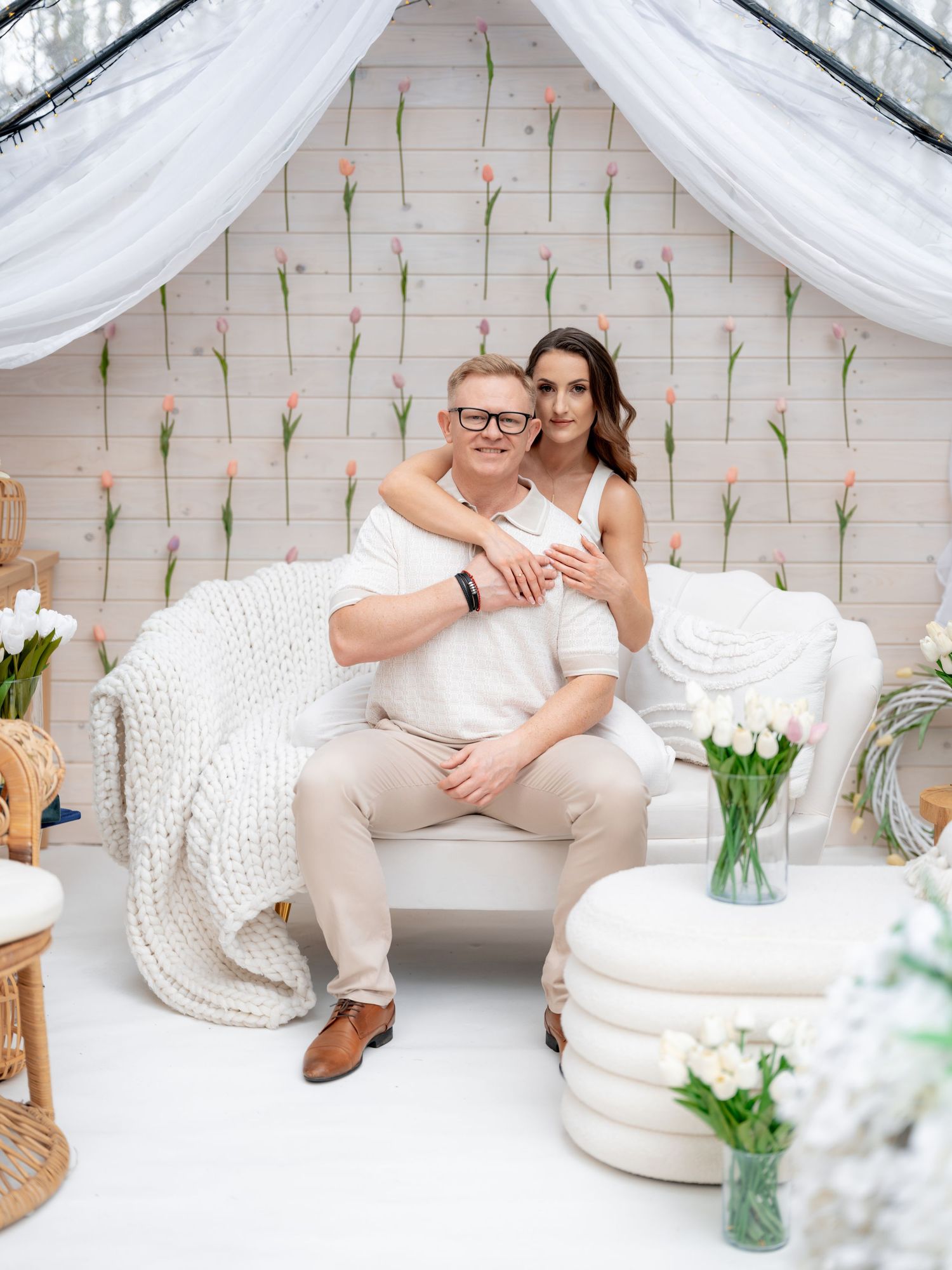 A couple relaxes on a white couch under flowing drapes with floral wall decorations.