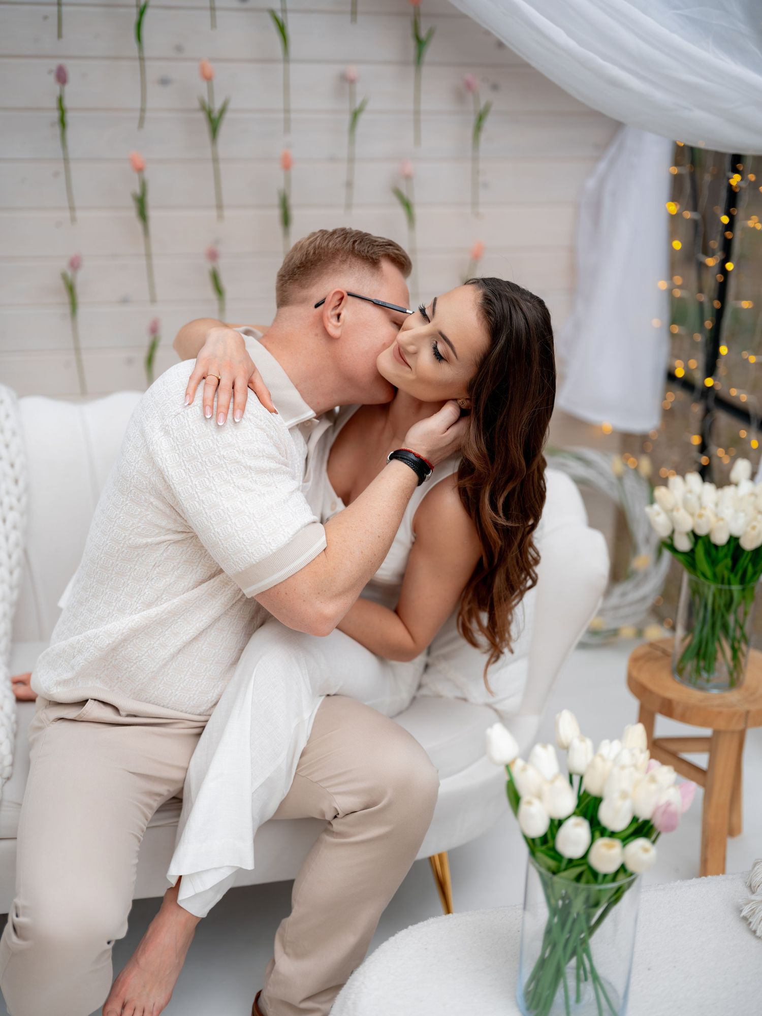 A romantic couple embraces on a white couch surrounded by white tulips and string lights in a dreamy setting.