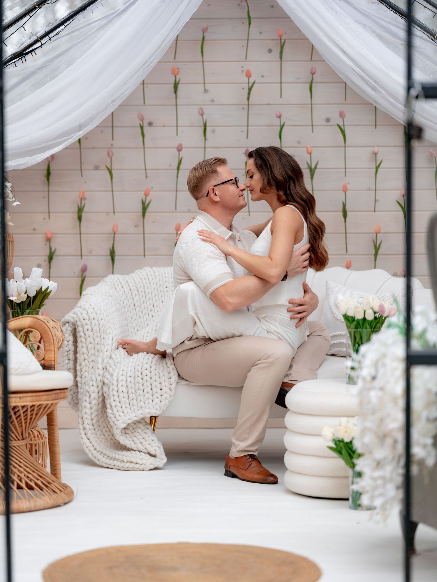 A romantic couple embraces on a white sofa with spring floral decor and white draping in an intimate setting.
