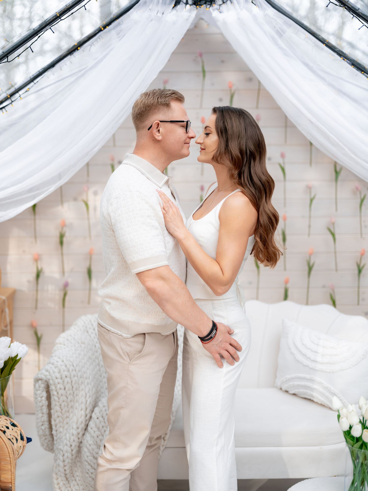 Romantic couple embraces under white draped canopy with floral backdrop and elegant decor.