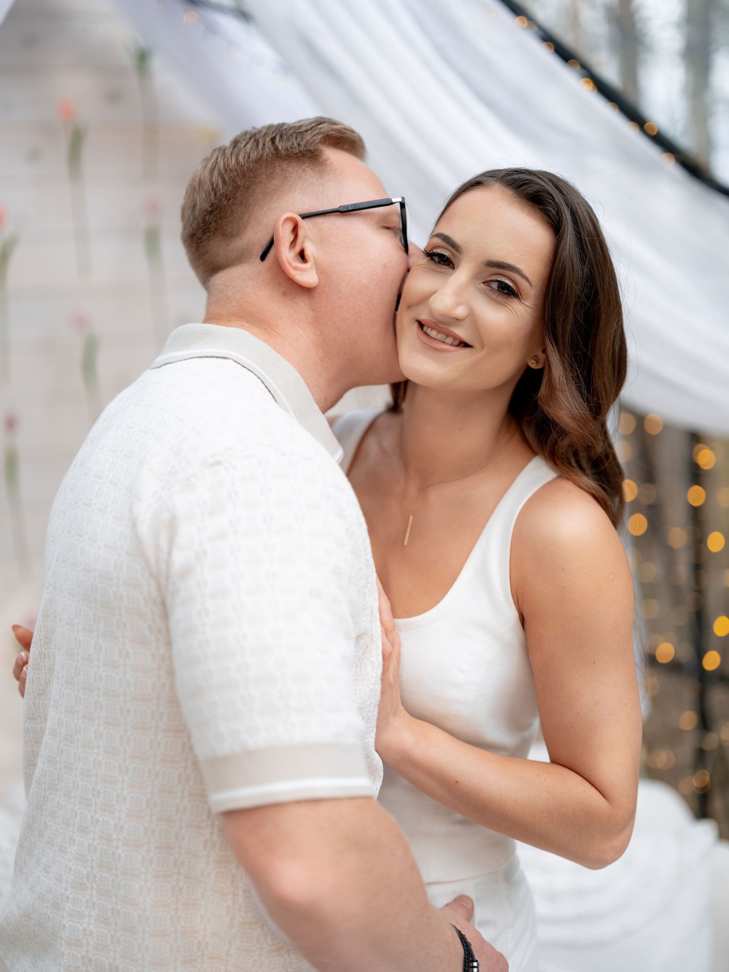 A romantic moment captured with a couple in white attire against a soft blurred background with twinkling lights.