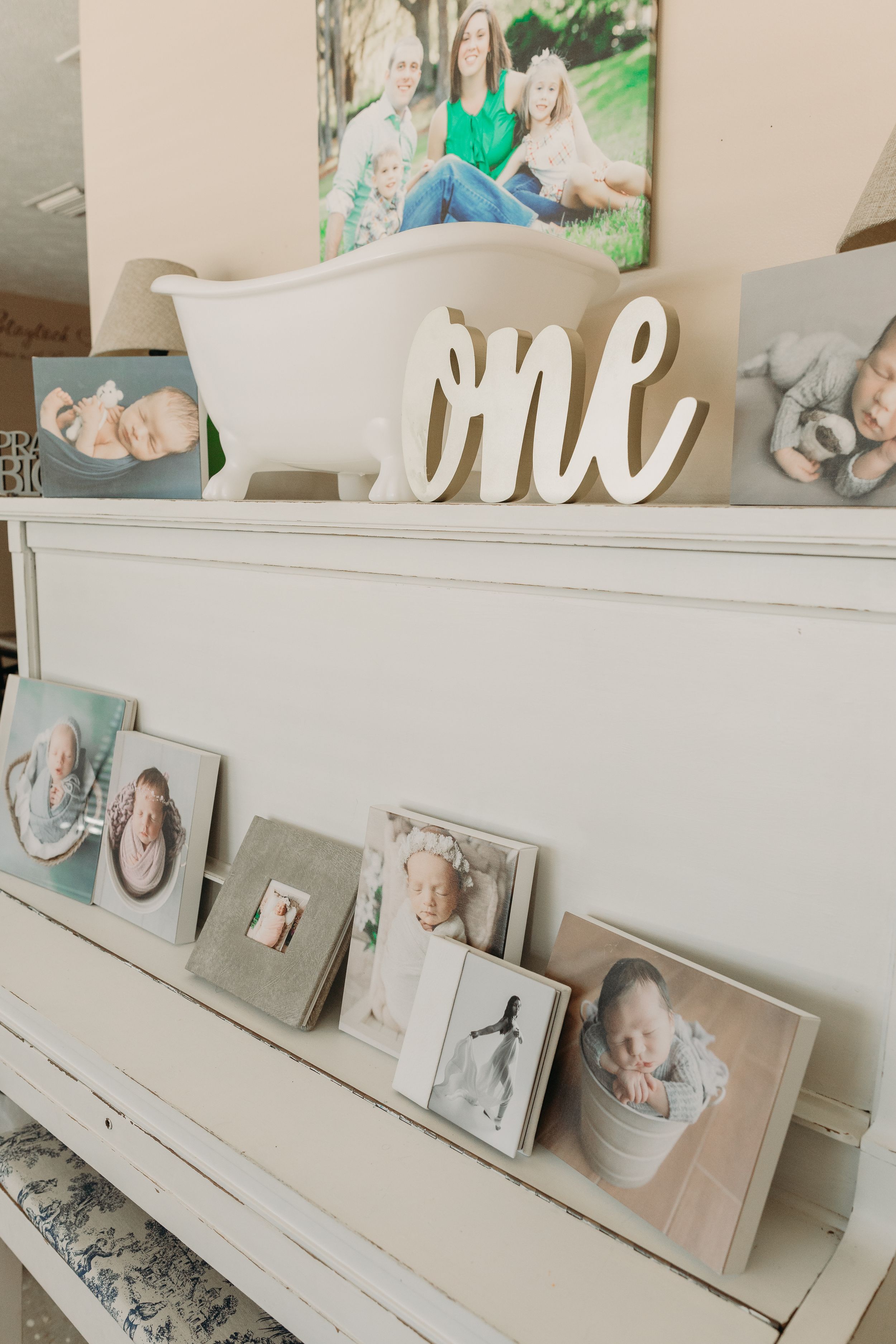 A white piano decorated with family photos and a wooden 'one' sign displayed on top features vintage-style picture frames.