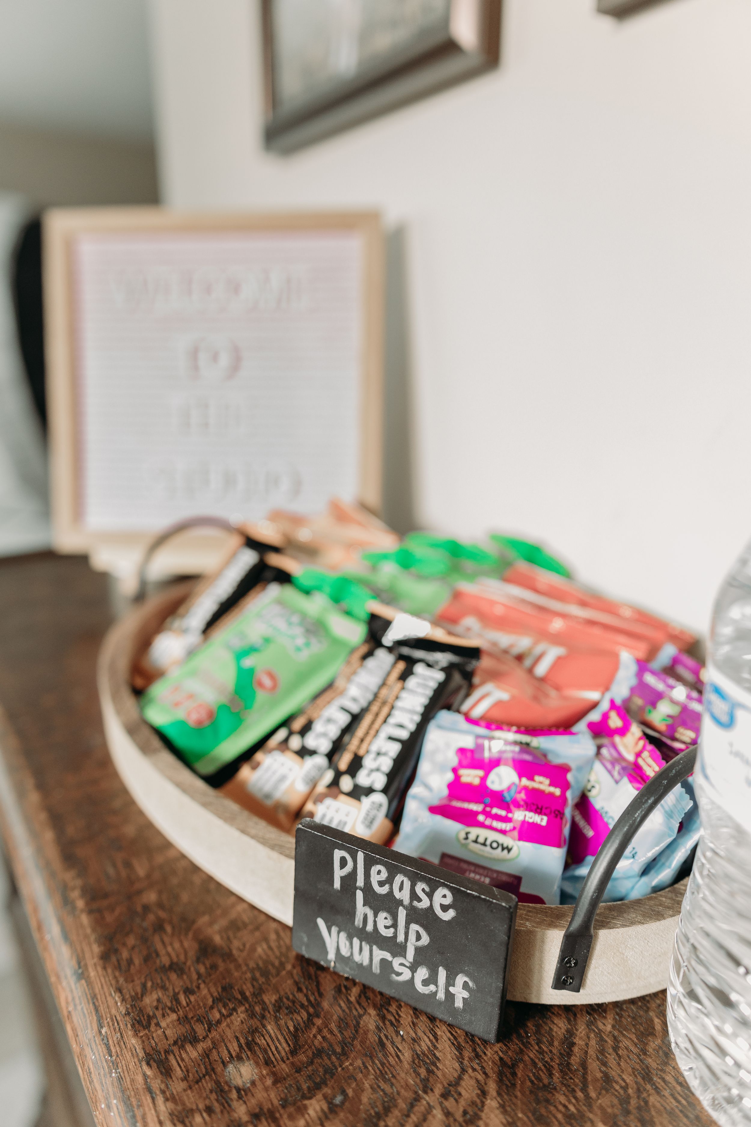 A wooden tray filled with assorted snacks and treats with a 'Please help yourself' sign on a dark wooden surface.