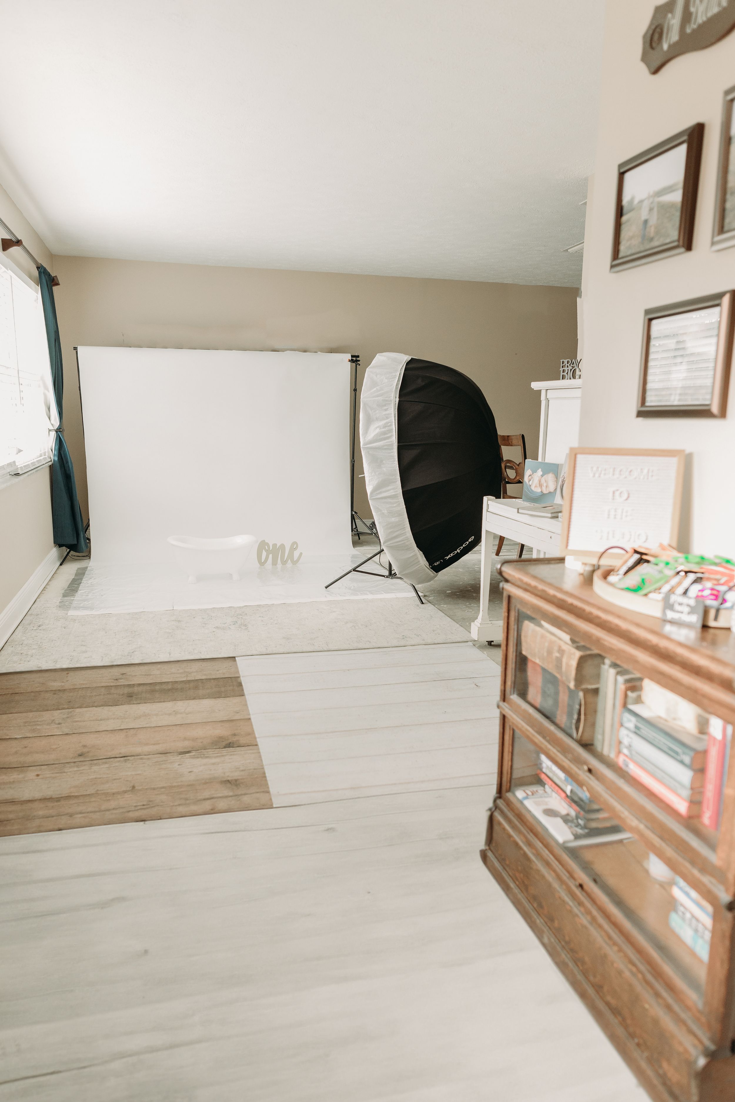 Photography studio setup with white backdrop, large softbox light, and wooden bookshelf storage in a home studio space.