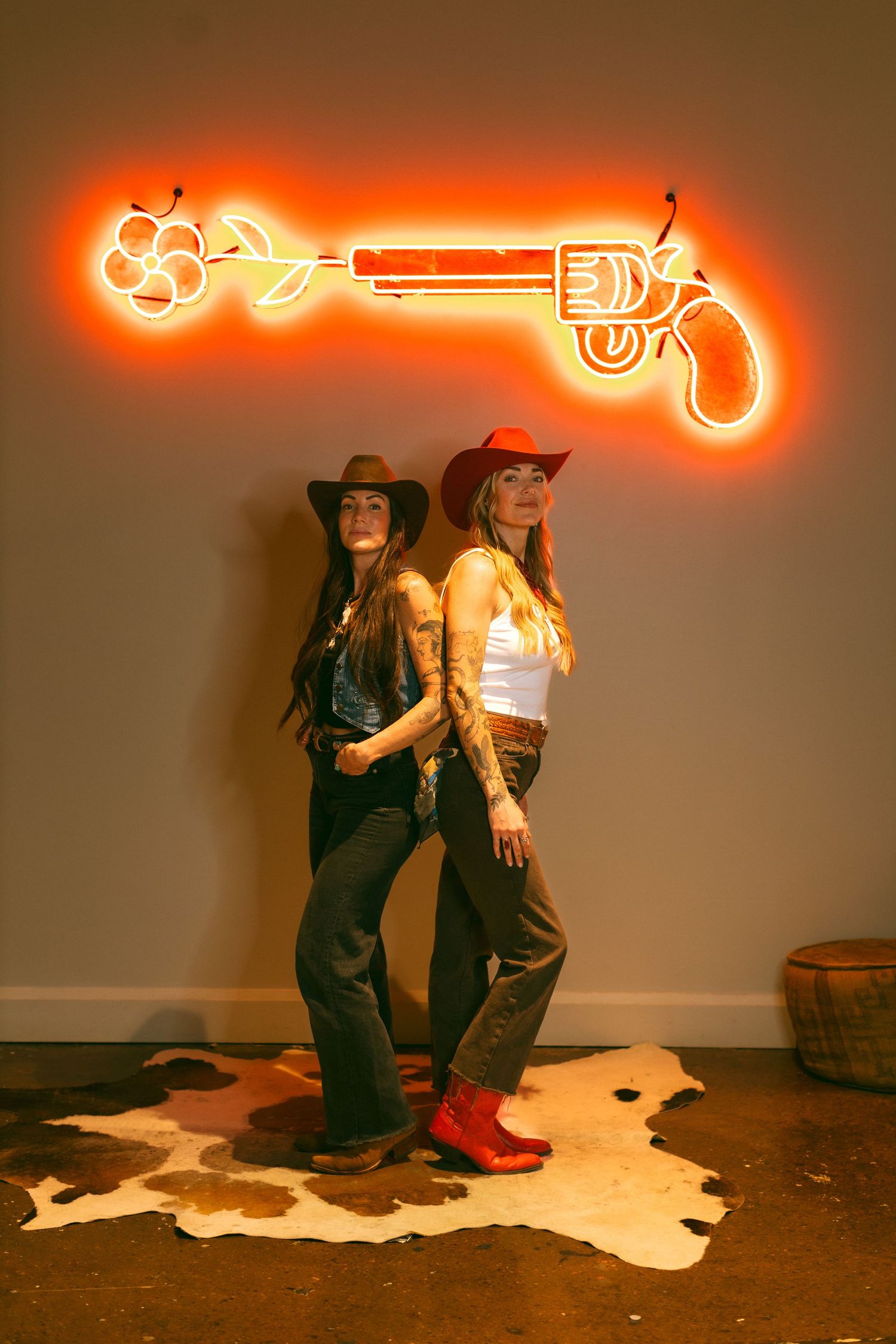 Two people in western wear pose against wall with glowing orange neon gun sign above on beige backdrop.