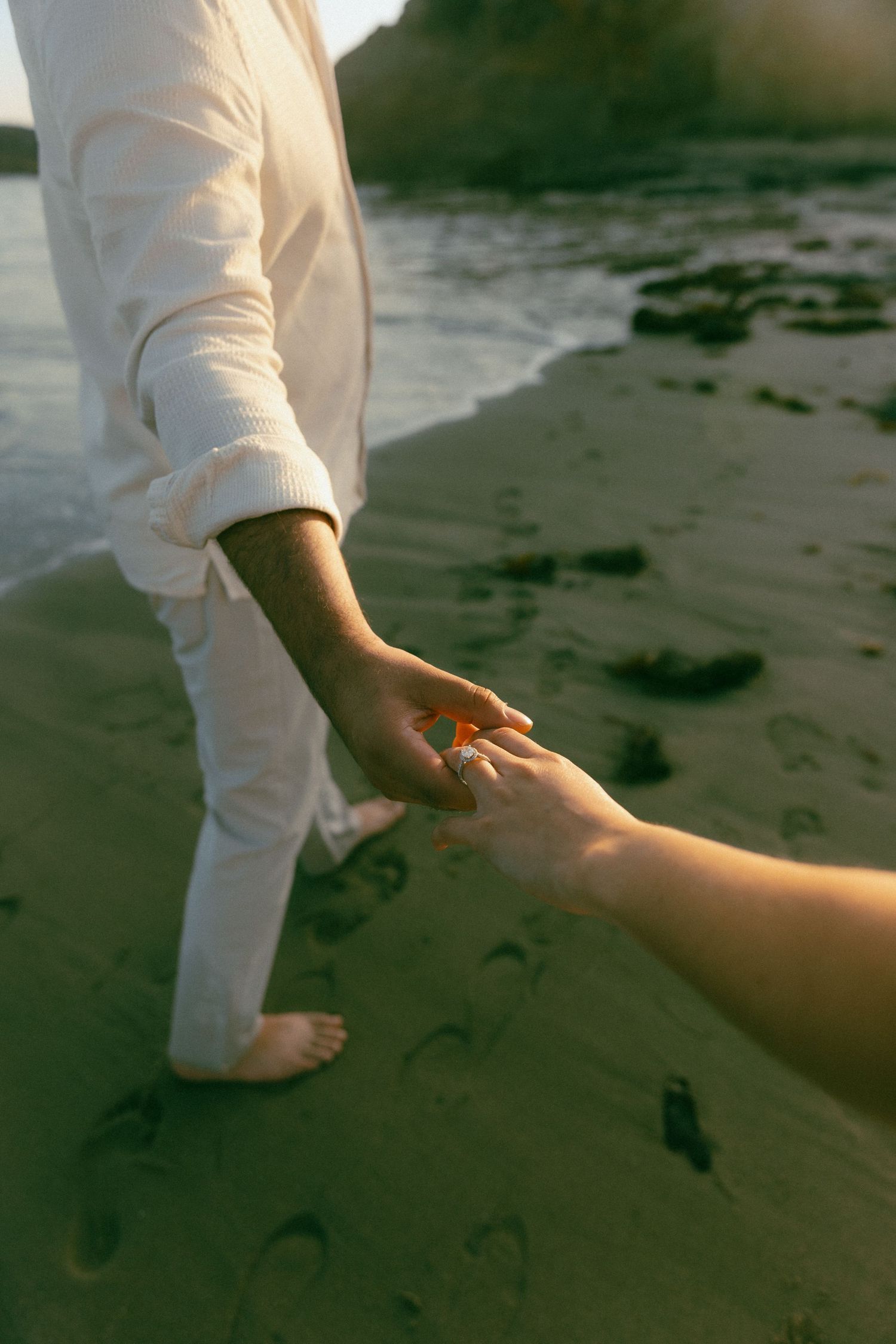 Two hands reaching towards each other on a sandy beach at sunset with one person wearing white clothing.