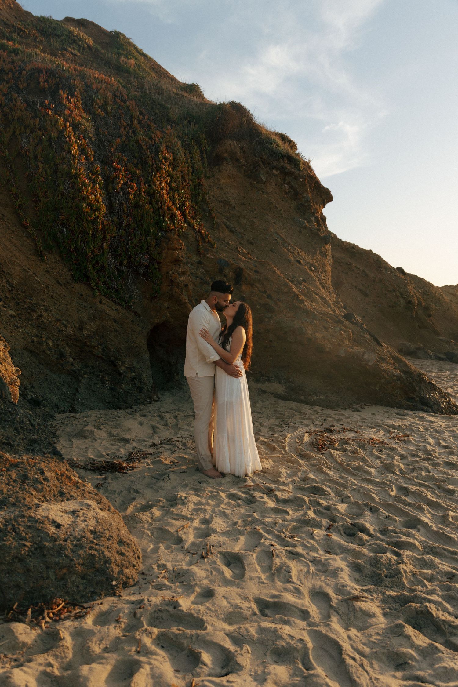 A couple in white outfits embrace on a sandy beach with dramatic cliffs during golden hour at sunset.