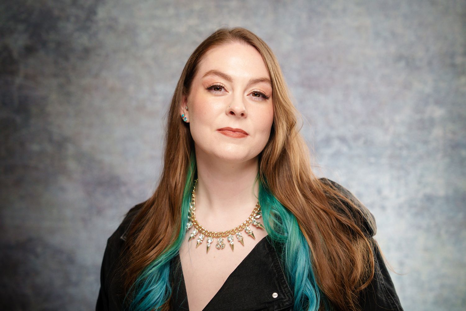 Studio portrait of someone with long blonde and teal-dipped hair wearing a black outfit and gold necklace against grey backdrop.