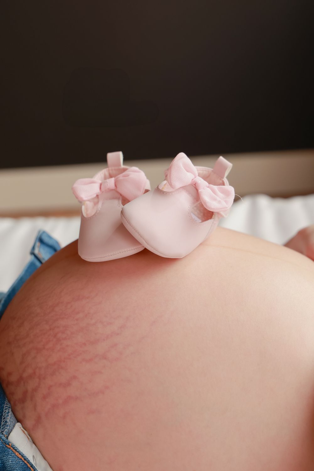 Pink baby shoes resting on pregnant belly against dark background.