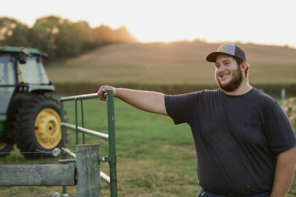 A farmer leans against farm equipment in a field at sunset with a John Deere tractor visible in the background.