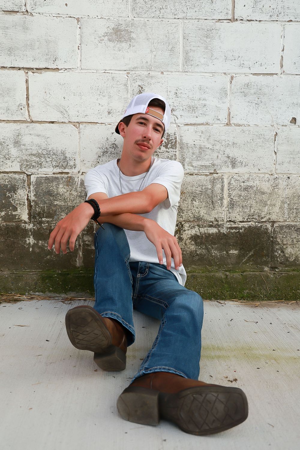 Person in white t-shirt and baseball cap lounges against concrete wall in casual pose.