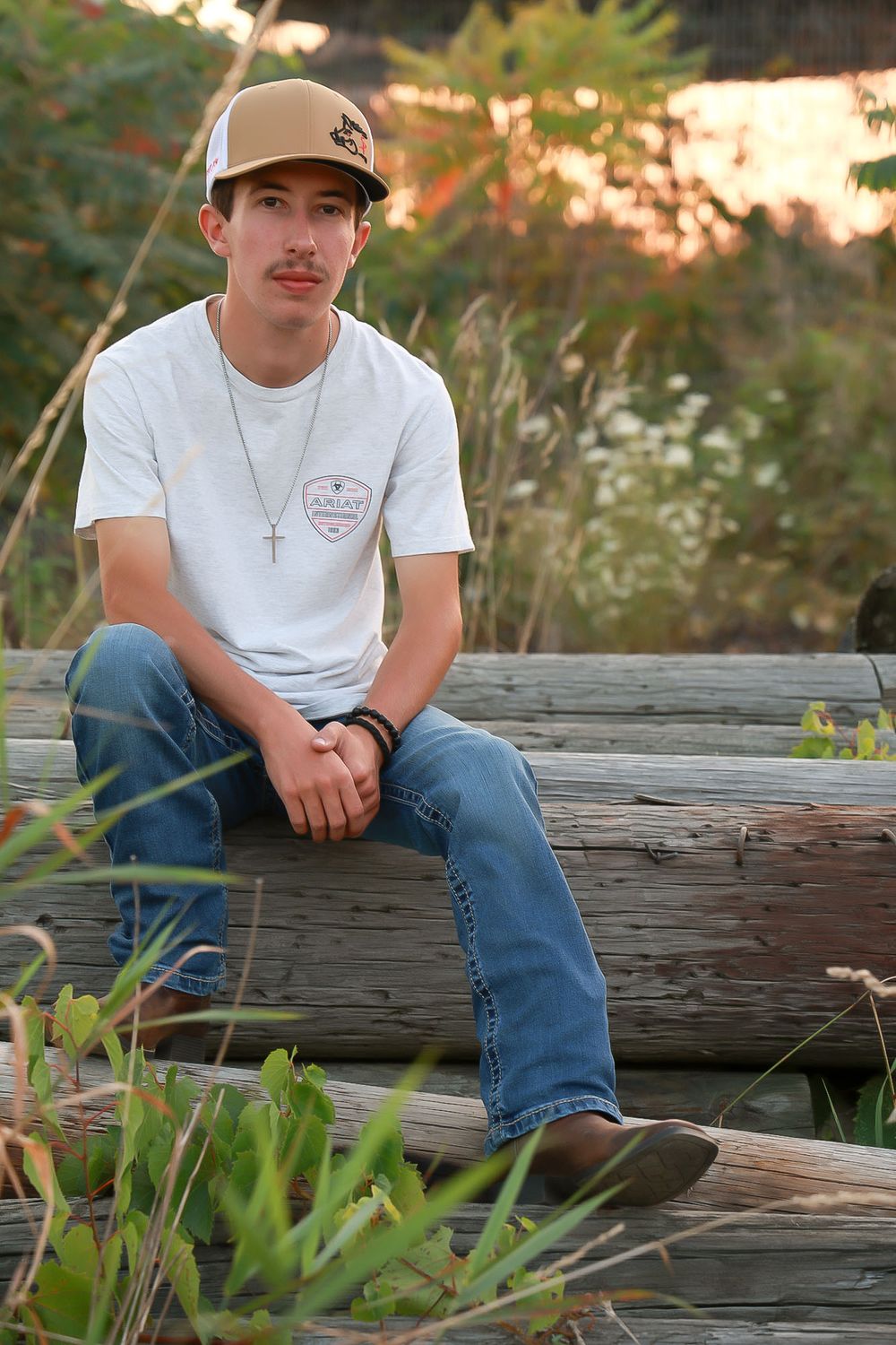 Person in baseball cap and white t-shirt sits on wooden log at sunset amongst grass.