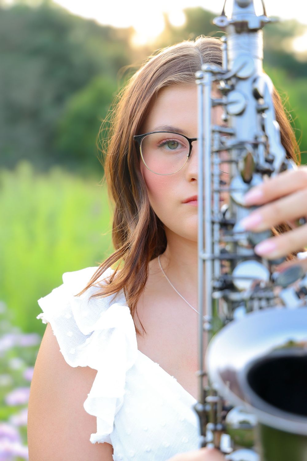 Person in white top poses with saxophone against blurred natural background.