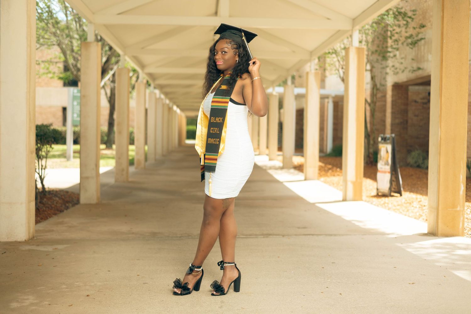 Graduate in white dress, cap, and kente stole poses under pergola columns for outdoor graduation portrait.