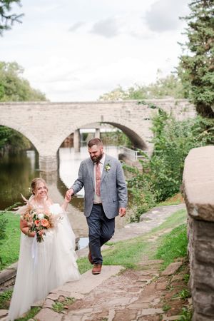 Bride and groom walk hand in hand along stone path beside river and arched bridge.
