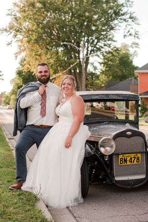 Smiling bride and bearded groom pose beside vintage black classic car on sunny street.