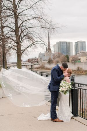 Groom dips bride for kiss as her lace veil billows dramatically in wind by waterfront.