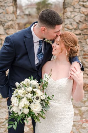 Groom kisses redhead bride on forehead as she smiles holding white floral bouquet.