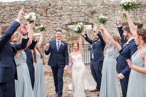 Bride and groom walk between cheering wedding party raising bouquets at stone venue.