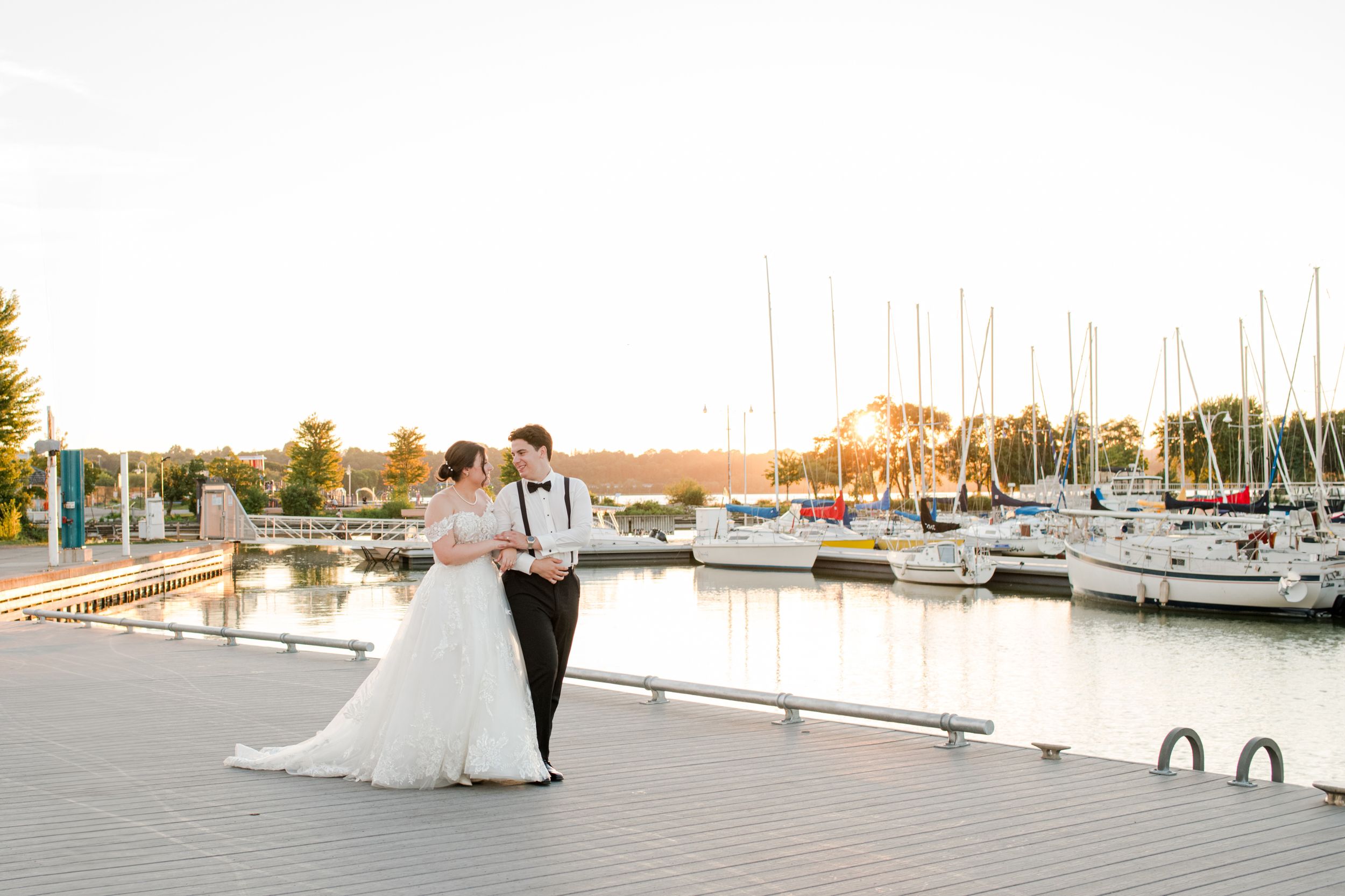 Bride and groom share romantic moment on marina dock at golden sunset with sailboats.