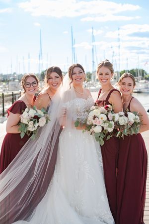 Bride in lace gown with veil poses with four bridesmaids in burgundy dresses holding white floral bouquets at a marina.