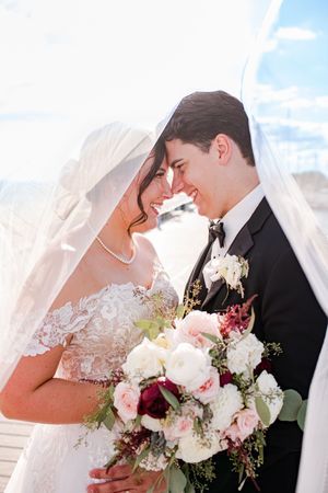 Bride and groom share an intimate moment under a veil, holding a bouquet of white and burgundy flowers.